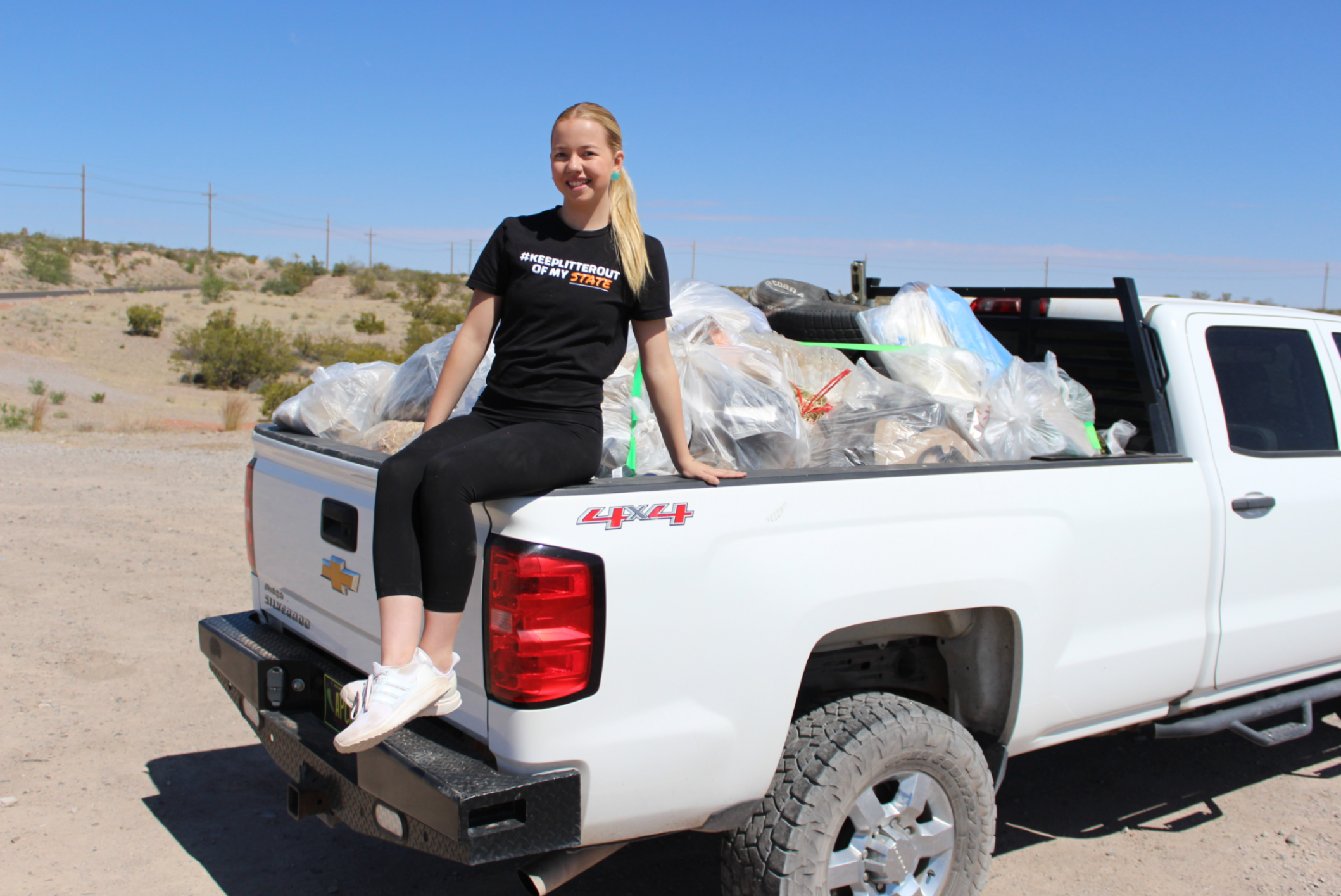 Young volunteer poses on a pickup truck filled with bags of collected litter following a community cleanup event in a New Mexico desert landscape.